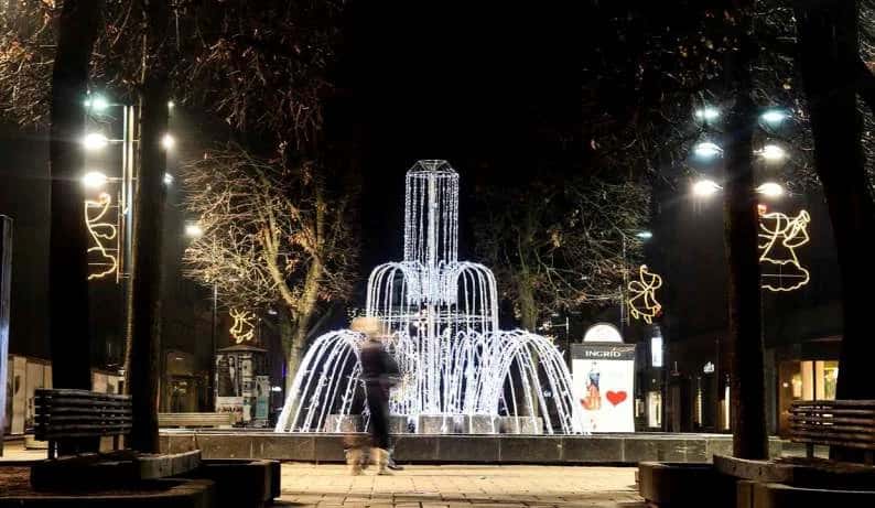 Kentsel meydanda ağaçlar arasında dev beyaz ışıklı merkez sütunlu fıskiye heykeli, gece