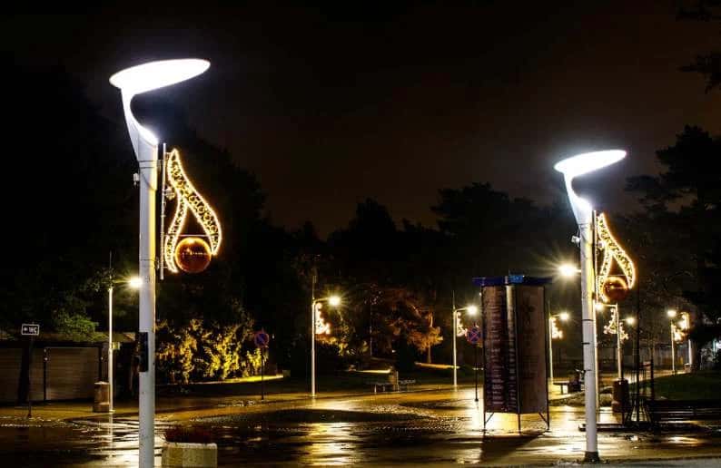 Park girişindeki iki lamba direğinde alev formunda LED motif ve altın küre, yağmurlu gece