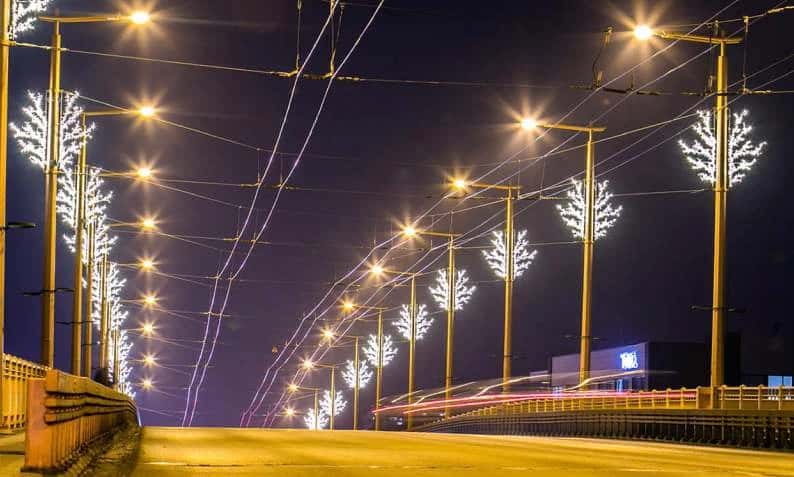 Köprü üzerinde tramvay direklerinin tepesine sıra halinde monte edilmiş beyaz ışıklı dal ağaçları, gece